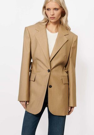 Blond naine, kellel on seljas suurepärase lõikega beež jakk, valge särk ja sinised teksad, seisab valge taustaga ees.