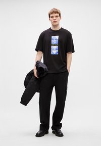 Μαύρο βαμβακερό T-shirt με μπλε και πράσινο γραφικό στο μπροστινό μέρος, συνδυασμένο με μαύρες cargo παντελόνες και κρατώντας μια μαύρη ζακέτα. Λείος υφασμάτινος καμβάς.