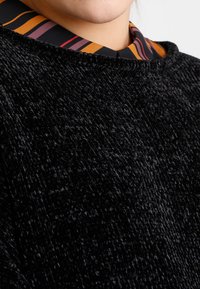 Black textured fabric of a top, featuring a contrasting striped collar in orange, burgundy, and black. Close-up view of material and details.
