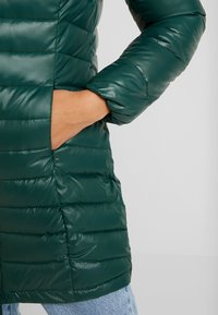 Abrigo acolchado verde con textura de acolchado, corte ajustado y bolsillos laterales. La tela parece brillante y suave con una longitud casual.