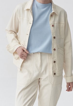 Veste en denim beige avec des boutons marron, portée sur un t-shirt bleu clair. Le mannequin porte également un pantalon beige assorti avec des poches.