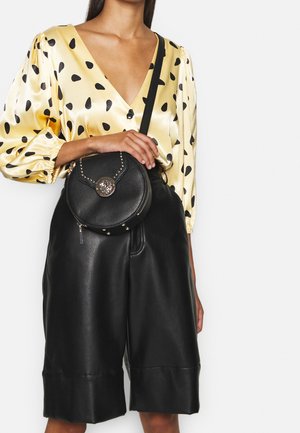 Kobieta w żółtej bluzce w groszki, czarnych skórzanych szortach do kolan, trzymająca czarną okrągłą torebkę na ramię z ozdobami w postaci złotych ćwieków.