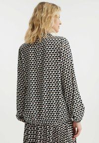 Zwarte en crèmekleurige blouse met een patroon en lange mouwen, voorzien van een elastische onderrand en een herhalend geometrisch ontwerp. Gladde stoftextuur.