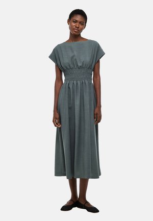 Femme debout portant une robe midi ample grise à manches courtes et taille haute, avec des chaussures plates noires sur un fond uni clair.