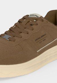 Καφέ sneaker Jack & Jones με υφασμάτινη υφή, κορδόνια και κρεμ χρώματος σόλα, που δείχνει το όνομα της μάρκας και "EST 1990" στην πλευρά.