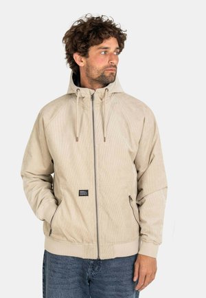 Mann mit lockigem Haar, der eine beige Kapuzenjacke mit Reißverschluss und blaue Jeans trägt, die Hände teilweise in den Jackentaschen, blickt nach rechts.