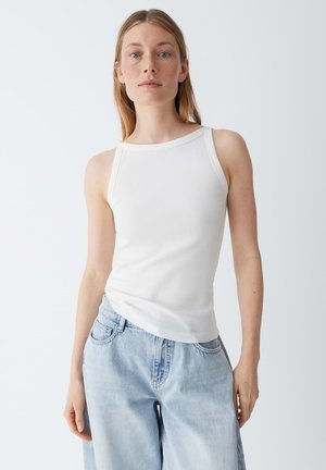 Femme portant un top blanc sans manches côtelé et un jean taille haute bleu clair, debout devant un fond clair uni.