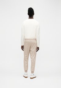 Tume näoga inimene, kellel on tume nahk ja punutud juuksed, kannab valget pika varrukaga särki, beežide mustriliste jalgpükse, valgeid sokke ja valgeid tossusid, seisab seljaga meie poole.