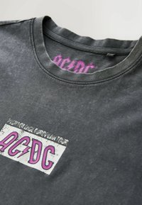 T-shirt de groupe AC/DC gris foncé avec un logo rose et blanc usé imprimé sur le devant et une étiquette de logo rose à l'intérieur du col.