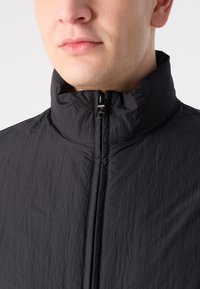 Jachetă ușoară neagră cu guler înalt, având un fermoar frontal și un material texturat, crăpat. Design minimalist fără modele vizibile.