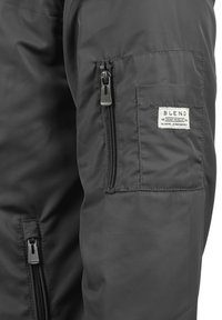 Veste isolante noire dotée d'une poche zippée sur la manche, d'une petite étiquette cousue et d'une texture de tissu lisse et brillante.