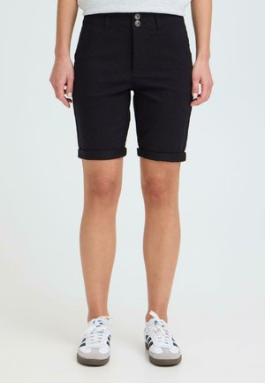 Person trägt schwarze Shorts mit umgeschlagenem Saum und weiße Turnschuhe mit schwarzen Streifen, steht vor einem schlichten hellen Hintergrund.