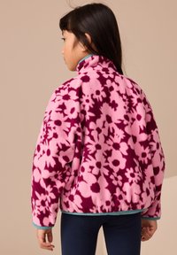 Rózsaszín polárdzseki sötét virágmintával. Magas gallérral és kontrasztos kék szegéllyel. Puha tapintású, laza fazon.