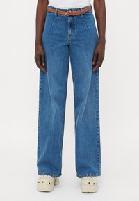 Jeans blu a gamba larga realizzati in denim, con vita alta, design a cinque tasche e una cintura in pelle marrone con fibbia argentata.