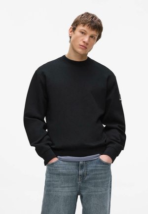Giovane uomo con una felpa nera semplice e jeans blu, in piedi con le mani nelle tasche davanti a uno sfondo bianco.