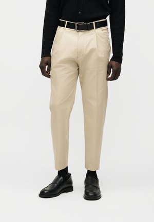 Bărbat purtând pantaloni plisati bej cu curea neagră, mocasini negri și bluză neagră cu mâneci lungi, stând în picioare pe un fundal alb simplu.