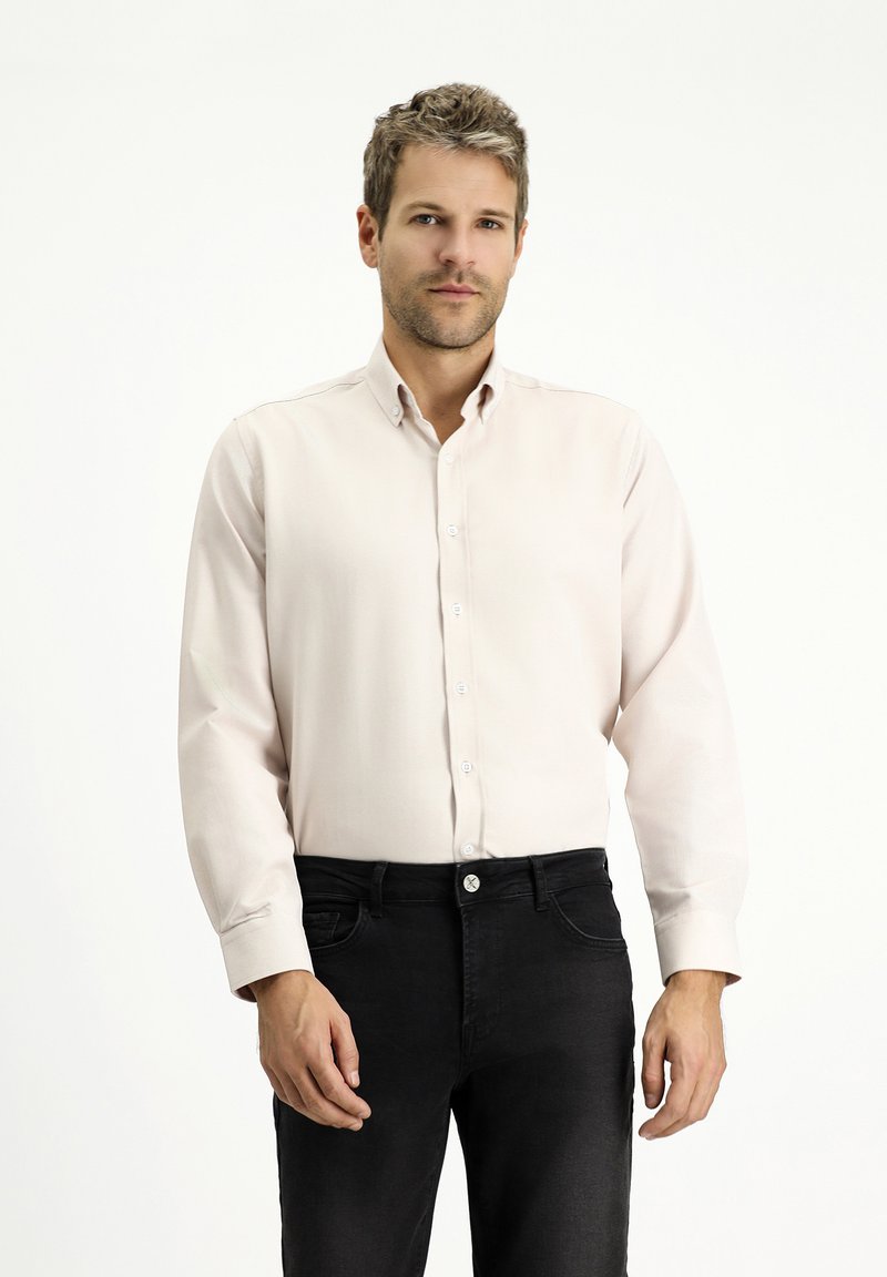 Camicia button-up beige chiaro con maniche lunghe, texture morbida e bottoni bianchi, abbinata a jeans neri. Design semplice e tagliato.