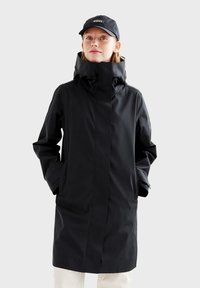 Veste noire imperméable avec col montant, fermeture éclair et boutons, poches latérales et capuche réglable. Portée avec une casquette noire.