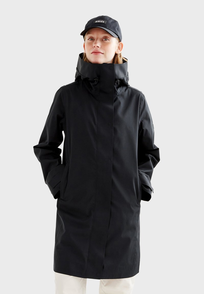 Veste noire imperméable avec col montant, fermeture éclair et boutons, poches latérales et capuche réglable. Portée avec une casquette noire.