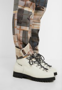 Bottes de randonnée en cuir blanc avec des lacets noirs et blancs, associées à un pantalon à carreaux beige et gris, maintenu par des brides de cheville beiges.