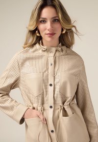 Beżowa perforowana kurtka z zapięciem na guziki, regulowany sznurek przy szyi, dwie przednie kieszenie oraz błyszczące akcenty w złotym kolorze.