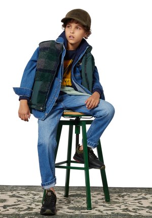 Veste en denim avec gilet en polaire verte, pantalon à motifs et chaussures noires. Le modèle est assis sur un tabouret vert devant un fond neutre.
