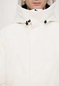 Veste isolante blanche avec un col montant, dotée de toggles noirs et de lignes épurées, fabriquée à partir d'un tissu lisse et texturé.