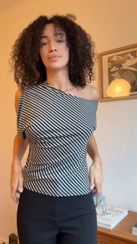 Melnbalts svītrainais tops ar asimetrisku izgriezumu un drapētiem auduma elementiem. Kombinēts ar melnām biksēm, kas izceļas ar gludu tekstūru un pieguļošu dizainu.