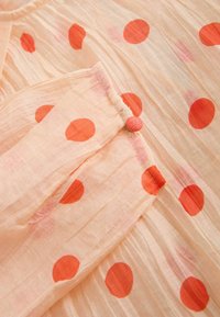 Vaalean persikanvärinen kangas, jossa isokokoisia oransseja pilkkuja, lähikuva napitetusta kalvosinnauhasta, jossa on teksturoitu, rypistynyt materiaali.