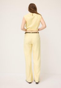 Femme debout, de dos à la caméra, portant un haut sans manches jaune pâle et un pantalon large assorti avec une ceinture marron.