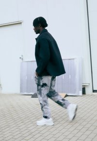 Fekete külső dzseki, világosszürke mintás nadrág sötét foltokkal, és fehér sportcipő. Sétál egy térkövezett felületen egy épület közelében.