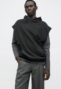 Nagy méretű fekete kapucnis pulóver, texturált felülettel és kapuccsal, szürke színű, testhez simuló hosszú ujjú póló fölött rétegezve, szürke nadrággal párosítva.