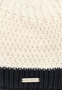 Stickad mössa i krämfärgad och marinblå med ett texturerat mönster. Silverfärgad logotyp på bandet. Mjukt garnmaterial.