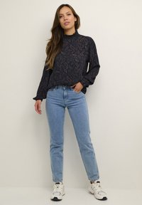 Tmavomodrá blúzka s paisley vzorom, vysokým volánovým golierom, dlhými rukávmi, v kombinácii so svetlomodrými džínsami a bielymi teniskami.