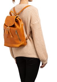 Zaino in pelle marrone chiaro con chiusura a pattina, lacci e due tasche laterali con zip. Design testurizzato e spallacci regolabili. Indossato su un maglione lavorato a maglia.