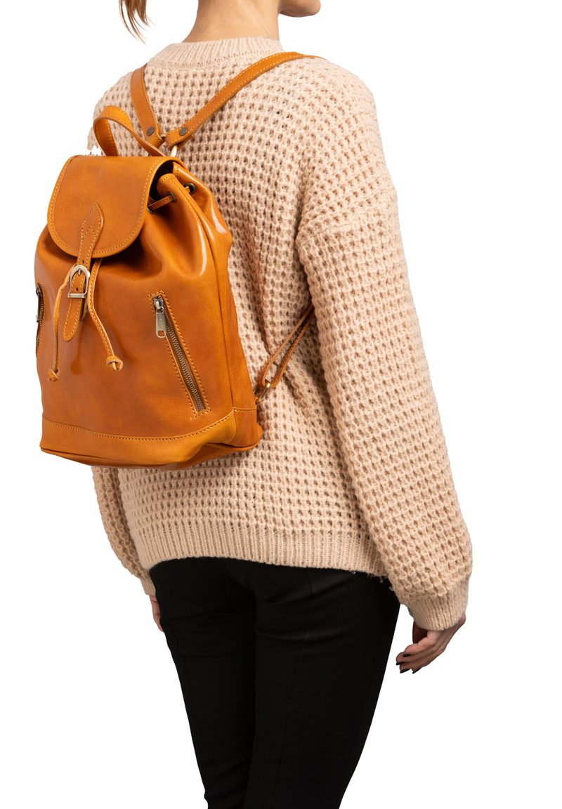 Zaino in pelle marrone chiaro con chiusura a pattina, lacci e due tasche laterali con zip. Design testurizzato e spallacci regolabili. Indossato su un maglione lavorato a maglia.