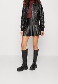 Svart läderjacka med dragkedja, skräddarsydd plisserad läderkjol och knähöga svarta stövlar med kraftiga sulor. Slät textur och skräddarsydd design.