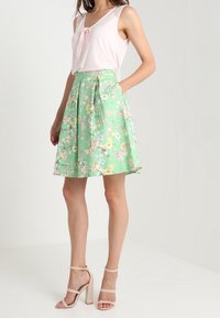 Haut rose clair sans manches avec un détail nœud, associé à une jupe verte à motifs floraux. Le mannequin porte des sandales à talons hauts couleur chair.