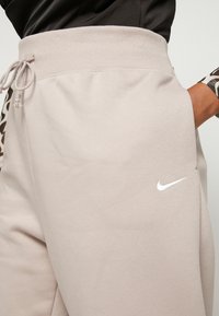 Svetlo bež trenerke z rebrastim elastikom v pasu, vrvico in majhnim belim logotipom Nike na strani. Mehka, teksturirana tkanina.