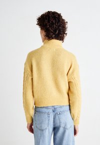 Pull tricoté jaune avec col haut, manches à torsades, ourlet côtelé et coupe décontractée, associé à un jean bleu clair.