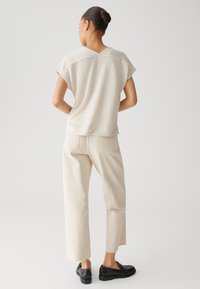 Top beige con texture, maniche a cappotto e scollatura a V, abbinato a pantaloni dritti color crema. Scarpe nere slip-on completano l'outfit.
