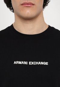Svart t-skjorte laget av bomull, med "ARMANI EXCHANGE" i tykk hvit skrift over brystet, rund halslinning og korte ermer.