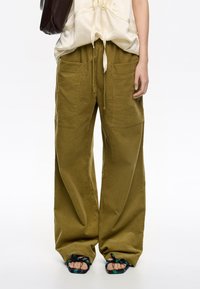 Pantalones de pierna ancha verde oliva con una tela texturizada y ligera; cuentan con dos bolsillos frontales y una cintura con cordón. Llevados con sandalias decorativas.