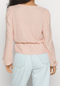 Ljusrosa strukturerad blus med långa puffärmar och figursydd midja, tillsammans med ljusblå högmidjade jeans. Bakvy visas.