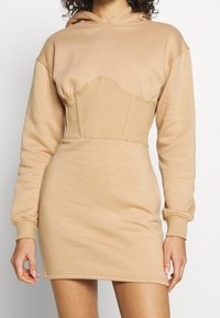 Vestido beige con capucha, detalles ajustados en el torso, mangas largas y textura suave. Presenta una sección acanalada en la cintura y el dobladillo.