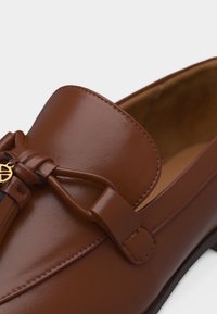 Καφέ δερμάτινο μοκασίνι με δεμένη λωρίδα και μικρό χρυσό στρογγυλό κρεμαστό στο πάνω μέρος, με ομαλή επιφάνεια και εμφανείς ραφές.