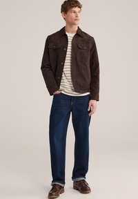 Ung mand stående iført en brun ruskindsjakke, stribet hvid og brun skjorte, mørkeblå jeans og brune læder snørestøvler.