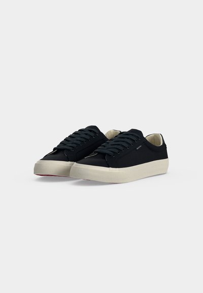 Paire de baskets basses noires avec lacets noirs et semelles épaisses blanc cassé sur un fond clair uni.