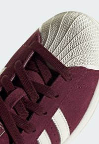 Burgundy suede sneaker with white leather stripes, featuring textured rubber toe cap and smooth laces. Close-up focus on materials and design.