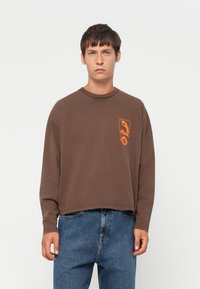 Mand iført en brun langærmet skjorte med en orange grafik på brystet og løstsiddende blå jeans, står op mod en ensfarvet baggrund.